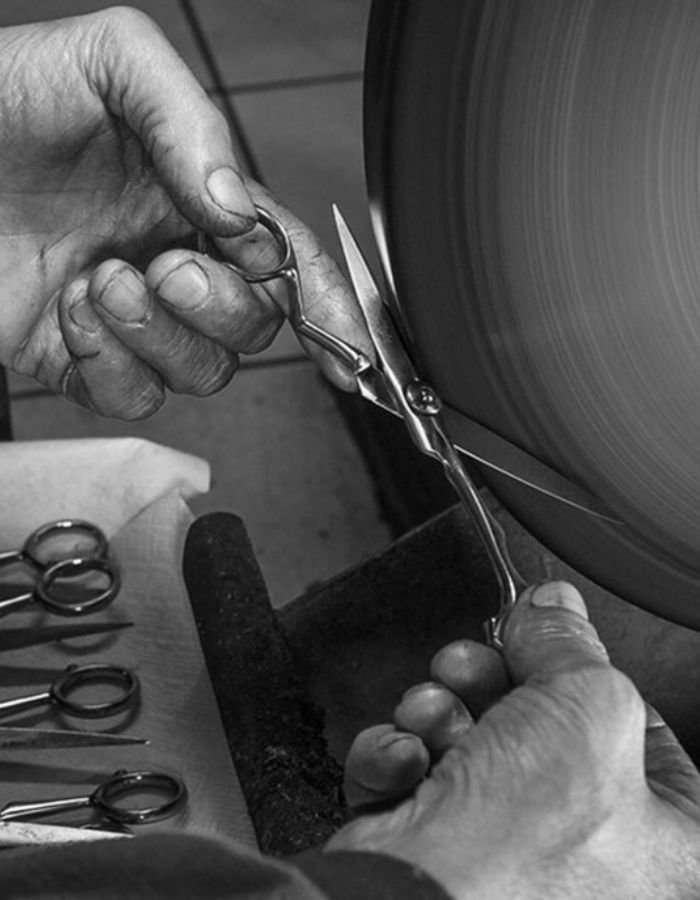 A skilled artisan’s hands meticulously guiding metal barber shears on a grinding wheel for professional scissor sharpening service in a workshop.