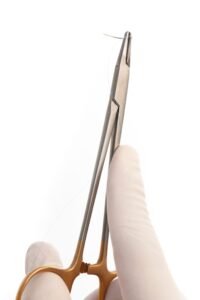 Surgical gloved hand demonstrating a gold-handle needle holder clamping a curved suture needle and thread.